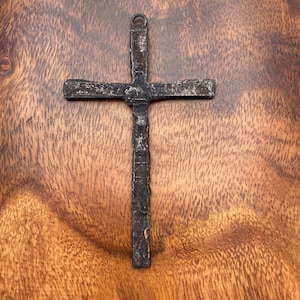May include: A dark metal cross with a textured surface and a loop at the top for hanging. The cross is set against a warm, wooden background, highlighting its rustic appearance.