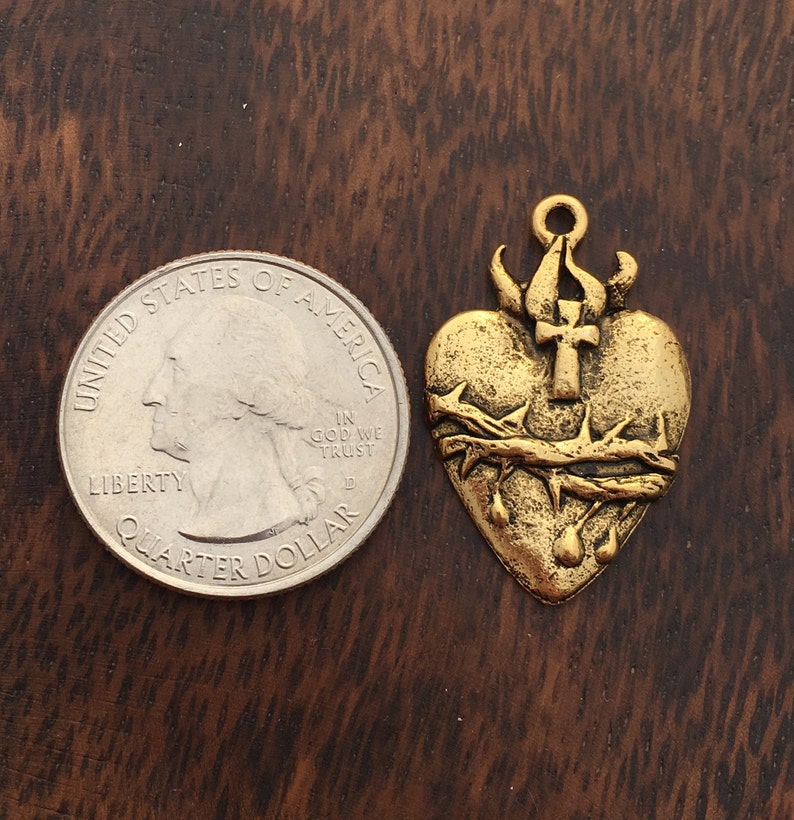 Op de afbeelding: Goudkleurige metalen hanger in de vorm van een hart met een doornenkroon, een kruis en bloeddruppels. De hanger is ongeveer zo groot als een Nederlandse 2 euro munt.