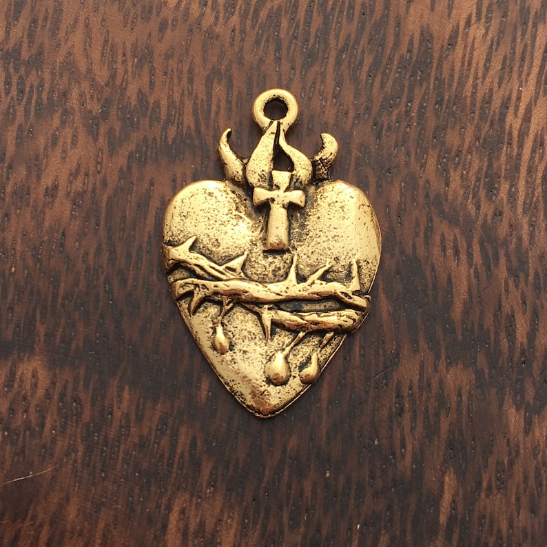 Op de afbeelding: Gouden metalen hanger in de vorm van een hart met een doornenkroon en een kruis erop. Het hart heeft een verweerde afwerking en lijkt te druppelen met bloed.