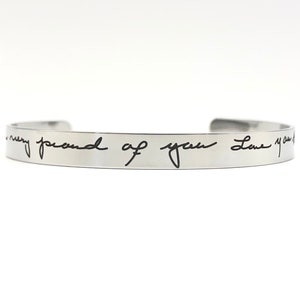 May include: Silver bangle bracelet with the inscription "I am very proud of you Love you" in black handwriting.