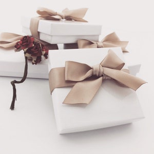 May include: Three white gift boxes with beige satin ribbon bows. The boxes are stacked on top of each other. The top box has a single bow, the middle box has a bow with a small bouquet of red flowers, and the bottom box has a large bow.