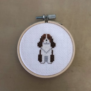 May include: A white cross-stitch embroidery of a brown and white dog in a wooden hoop.