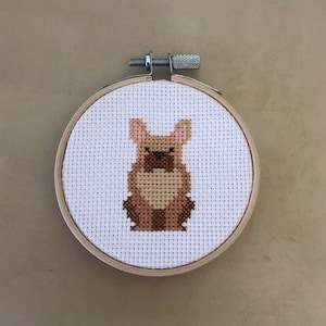 May include: A brown and white cross-stitch embroidery of a French bulldog in a wooden embroidery hoop.