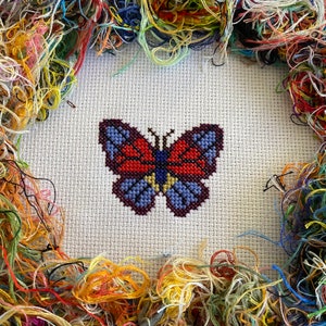 Puede incluir: Un primer plano de un bordado de punto de cruz de una mariposa con acentos rojos, naranjas, azules y amarillos. La mariposa está rodeada por un borde de hilo de bordar de colores.