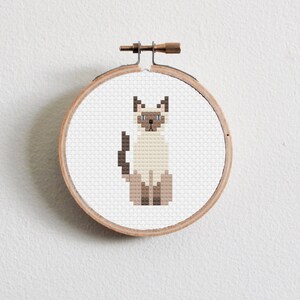 May include: A cross-stitch embroidery of a Siamese cat in a wooden hoop. The cat is depicted in shades of beige, brown, and black, with blue eyes. The embroidery is set against a white background.