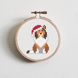 May include: A cross-stitch pattern of a dog wearing a red Santa hat. The dog is brown and white with a gray nose and black eyes. The pattern is framed in a round wooden hoop.