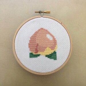 Puede incluir: Un aro de bordado de punto de cruz con un diseño de melocotón. El melocotón es rosa y amarillo con hojas verdes.