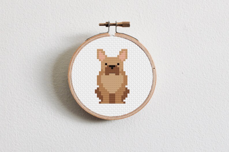 May include: A brown and white cross-stitch pattern of a French bulldog in a wooden embroidery hoop.