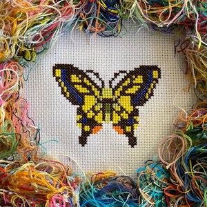 Puede incluir: Un patrón de mariposa amarilla y azul en punto de cruz sobre tela blanca, rodeado de hilo de bordar de colores.