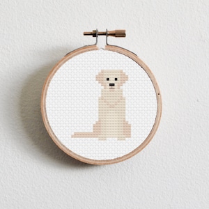 Könnte beinhalten: Ein Kreuzstich-Stickrahmen mit weißem Hintergrund und einem beigefarbenen Hund, der in der Mitte sitzt. Der Hund hat eine braune Nase und schwarze Augen.
