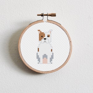 Puede incluir: Una bordado de punto de cruz blanco, marrón y gris de un perro sentado en un aro de bordado de madera.