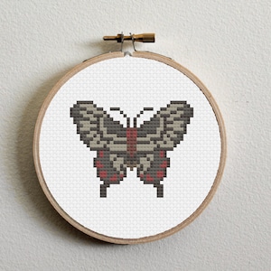 Puede incluir: Un aro de bordado de punto de cruz con una mariposa marrón y blanca con detalles rojos. La mariposa está centrada en el aro.