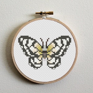 Puede incluir: Una mariposa blanca y negra con detalles amarillos está bordada en un lienzo blanco, enmarcada en un aro de madera.