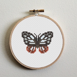 Puede incluir: Un aro de bordado blanco con un diseño de mariposa en punto de cruz. La mariposa es marrón, blanca y naranja.