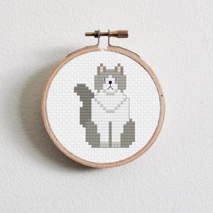 Puede incluir: Un patrón de punto de cruz de un gato gris y blanco en un aro de bordado de madera.