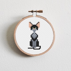Puede incluir: Un gato negro y gris con orejas de color melocotón está bordado en punto de cruz sobre tela blanca en un aro de bordado de madera redondo.