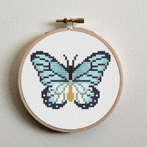 Puede incluir: Un aro de bordado con una obra de arte de bordado de cruz de mariposa azul y blanca.