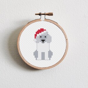 May include: A white dog wearing a red Santa hat, embroidered on a white fabric stretched over a wooden embroidery hoop.