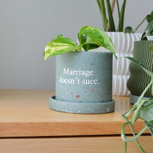 May include: A cylindrical sage green planter with a speckled terrazzo finish and a matching saucer. The front of the planter features the white printed text "Marriage doesn't succ." A small green and yellow variegated plant is potted inside the container.