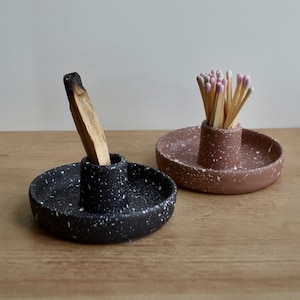 Puede incluir: Dos portavelas de cerámica moteados para incienso y cerillas. El porta incienso es negro con motas blancas y contiene un palo de incienso parcialmente quemado. El porta cerillas es rosa con motas blancas y contiene un paquete de cerillas con puntas rosas.