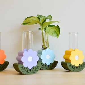 May include: Four colorful flower-shaped plant holders with green leaves. Each holder has a clear glass tube for a plant. The holders are in shades of orange, purple, blue, and yellow.
