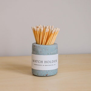 Puede incluir: Un porta-cerillas de hormigón azul claro con una etiqueta blanca que dice "MATCH HOLDER HANDMADE IN BROOKLYN, NY". El porta-cerillas está lleno de cerillas de madera con puntas blancas.