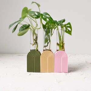 May include: Three glass test tubes with green plants are displayed in a set of three arched planters. The planters are green, yellow, and pink.