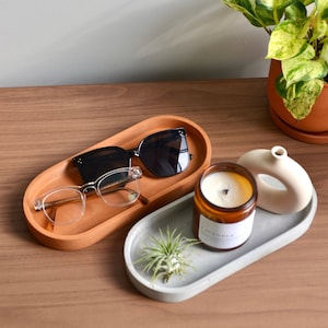 May include: Two oval-shaped trays, one terracotta and one gray concrete, with various items on top. The terracotta tray holds a pair of black sunglasses and clear glasses. The gray tray holds a small potted air plant, a brown candle with the word "Lavender" on the label, and a white ceramic sculpture.