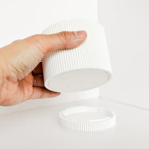 Könnte beinhalten: Ein weißer, zylindrischer Behälter mit geriffelter Außenseite und einem separaten, passenden Deckel. Der Behälter wird in einer Hand gehalten, wobei der Deckel darunter positioniert ist. Das Design ist minimalistisch und modern.