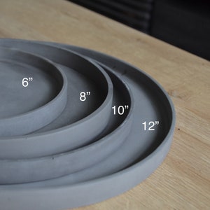 May include: Four gray concrete serving trays of varying sizes, 6", 8", 10", and 12" in diameter, are stacked on a light wood surface. The trays have a simple, modern design with a slightly raised edge.