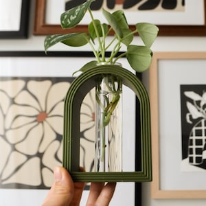 May include: A green ceramic arch vase with a clear glass tube for holding a plant. The plant is a green leafy plant with roots visible in the water.