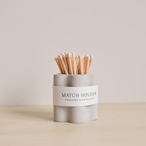 Puede incluir: Un porta fósforos de hormigón gris con un diseño ondulado. El porta fósforos tiene una etiqueta blanca que dice "MATCH HOLDER HANDMADE IN BROOKLYN, NY".