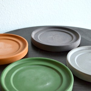 May include: Four round plant saucers in different colors: brown, gray, light gray, and green. The saucers are made of a textured material and are arranged on a black surface.