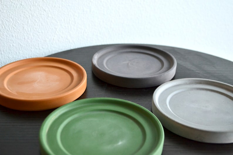 May include: Four round plant saucers in different colors: brown, gray, light gray, and green. The saucers are made of a textured material and are arranged on a black surface.