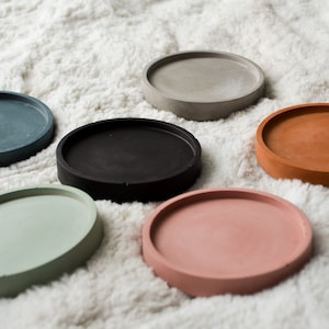 May include: Six round concrete coasters in various colours, including grey, black, green, and pink. The coasters are arranged on a white fluffy surface.