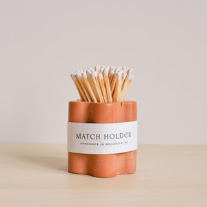 Puede incluir: Un portavelas de terracota con un diseño ondulado y una etiqueta blanca que dice "MATCH HOLDER HANDMADE IN BROOKLYN, NY". El portavelas está lleno de cerillas de madera con puntas blancas.