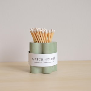 Puede incluir: Un porta cerillas de hormigón verde con una etiqueta blanca que dice "MATCH HOLDER HANDMADE IN BROOKLYN, NY". El porta cerillas está lleno de cerillas de madera con puntas blancas.