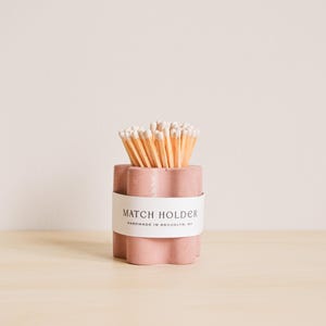 Puede incluir: Un porta-cerillas de hormigón rosa con una etiqueta blanca que dice "MATCH HOLDER HANDMADE IN BROOKLYN, NY". El porta-cerillas está lleno de cerillas de madera.