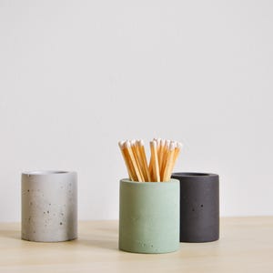 Puede incluir: Tres portavelas de hormigón, uno gris, uno verde y uno negro. El portavelas verde contiene cerillas.