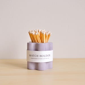Puede incluir: Un porta cerillas de hormigón de color lila claro con una etiqueta blanca que dice "MATCH HOLDER HANDMADE IN BROOKLYN, NY". El porta cerillas está lleno de cerillas de madera.