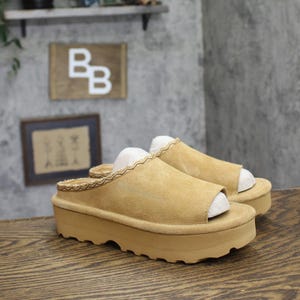 May include: A pair of tan suede platform slide sandals featuring an open-toe design and a thick, textured rubber sole. The upper edge of the sandal is finished with a decorative braided trim detail, providing a consistent look across the footwear.