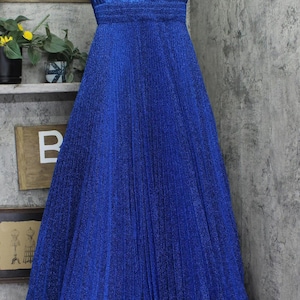 May include: A long, royal blue dress with a V-neckline and pleated skirt. The dress has a shimmering, textured fabric. The dress is hanging on a metal hanger against a gray wall.
