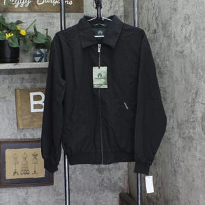 May include: A black jacket with a zipper closure, long sleeves, and a collar is hanging on a metal rack. The jacket has a tag attached with a logo and text. The background includes a shelf with plants and a framed picture.