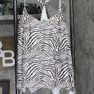 May include: A sleeveless top with a zebra print pattern in shades of brown and white. The top is hanging on a metal hanger. The straps cross in the back. The top has a V-neckline and a white lining.