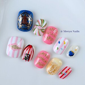 Puede incluir: Un juego de diez uñas postizas con diseños coloridos que incluyen una uña azul con una rana marrón, uñas rosas con el texto HAPPEE BIRTHDAE HARRY, uñas a rayas blancas y rosas, una uña con purpurina dorada y motivos de pequeños dulces.