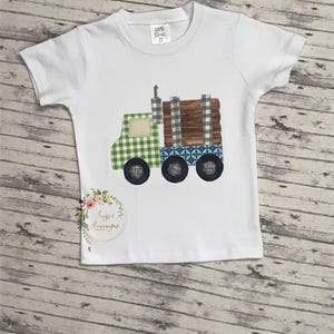 May include: A white short-sleeve t-shirt featuring a fabric applique of a logging truck. The truck cab is green gingham, the trailer is brown wood-grain fabric with grey plaid accents, and the wheels are black. A size 2T tag is at the neck.