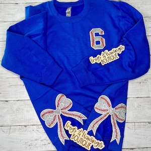 May include: A royal blue crewneck sweatshirt featuring a large silver glitter number 6 with red baseball stitching details on the chest. Two large silver glitter bows with red baseball stitching are positioned on the lower front of the garment.