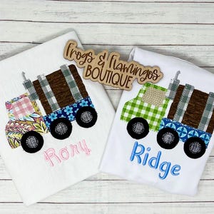 May include: Two white t-shirts featuring embroidered applique logging trucks. The left truck has pink and floral patterns with the name Rory in pink. The right truck has green gingham patterns with the name Ridge in blue. A wooden sign reads Frogs & Flamingos BOUTIQUE.