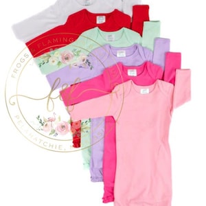 May include: A stack of long-sleeved baby gowns in various solid colors including white, red, light green, lavender, hot pink, and light pink. Each gown features a round neckline, long sleeves, and a gathered bottom hem, arranged in a cascading vertical row.