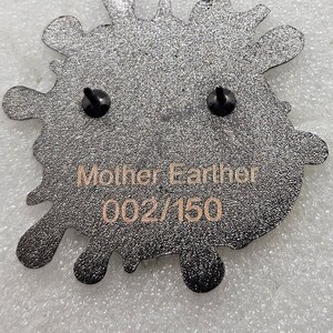 May include: Black enamel pin in the shape of a splatter with two black pin backs. The pin has the text "Mother Earth" and "002/150" in gold lettering.